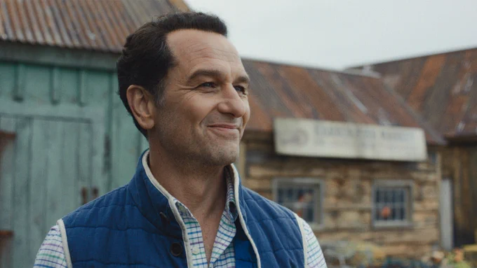 From Widow's Bay, a close-up shot of Matthew Rhys as Mayor Tom Loftis, smiling wearing a blue vest, standing in front of seaside buildings.