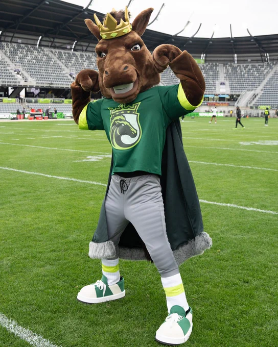 A horse mascot wearing a crown and green outfit poses energetically on a football field. The stadium in the background is partially filled.