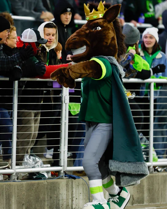 A lively sports mascot wearing a crown engages with fans at a game. The atmosphere is energetic and filled with excitement.