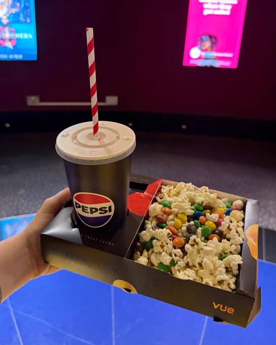A person holds a tray with a Pepsi cup featuring a striped straw, and a serving of popcorn mixed with colorful candy. In the background, there are two brightly lit movie posters inside a Vue cinema.