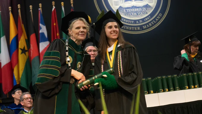 Graduation ceremony with President Julia Jasken awarding student Maria Gullo with her diploma on stage. There's a backdrop of international flags and the McDaniel College seal behind them.
