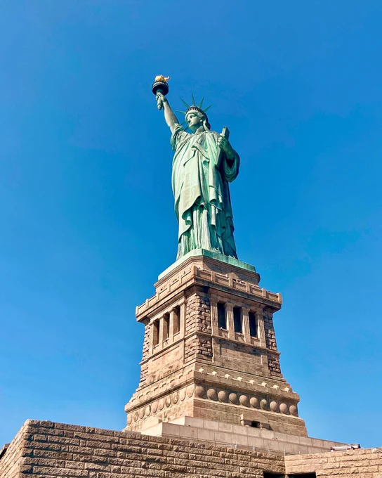 The image shows the Statue of Liberty against a clear blue sky. The statue is on a stone pedestal, and the iconic torch is held high in one hand.