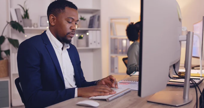 Man in a navy blazer typing on a keyboard at a modern office desk with a large monitor in front of him.