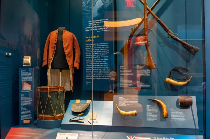 Display of New England soldier artifacts at the Museum of the American Revolution. Includes a coat, drum, gunpowder horns, and muskets. Informational text is visible in the background.