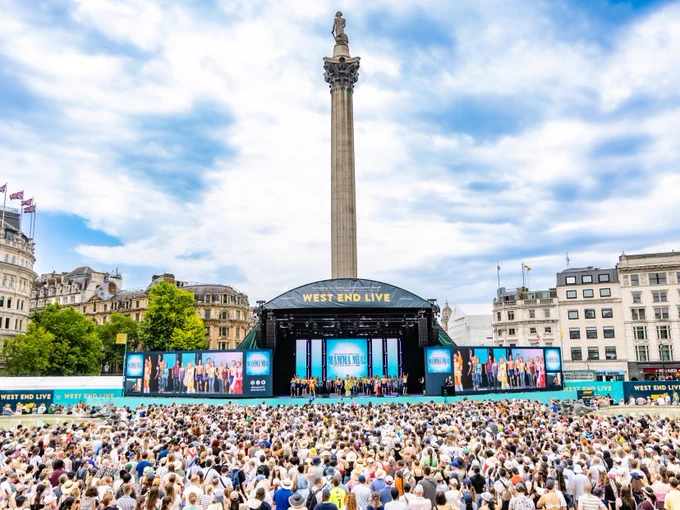 A large crowd is gathered at Trafalgar Square in London for a "West End Live" event. In the background, Nelson's Column rises prominently. Various performers are on stage with digital screens displaying the "West End Live" logo. Historic buildings surround the square under a partly cloudy sky.