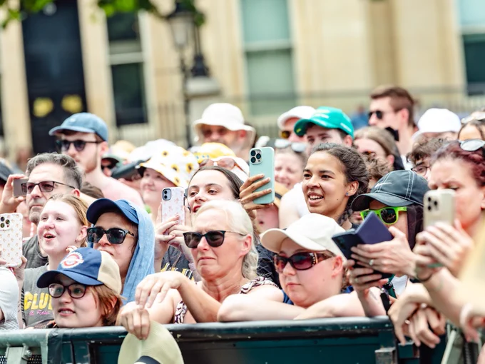 A diverse crowd of people gathers outdoors, holding smartphones to capture the event. Many are wearing sunglasses and hats. The atmosphere appears excited and engaged.