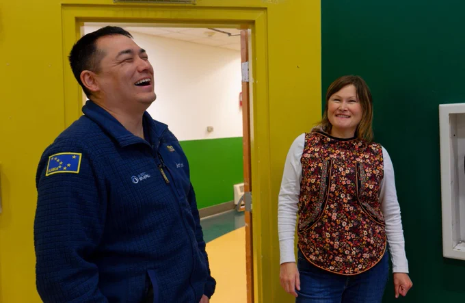 Two people are standing indoors near a yellow wall. One is wearing a jacket with a patch of the Alaskan flag. They are both smiling. The walls are painted green and yellow.